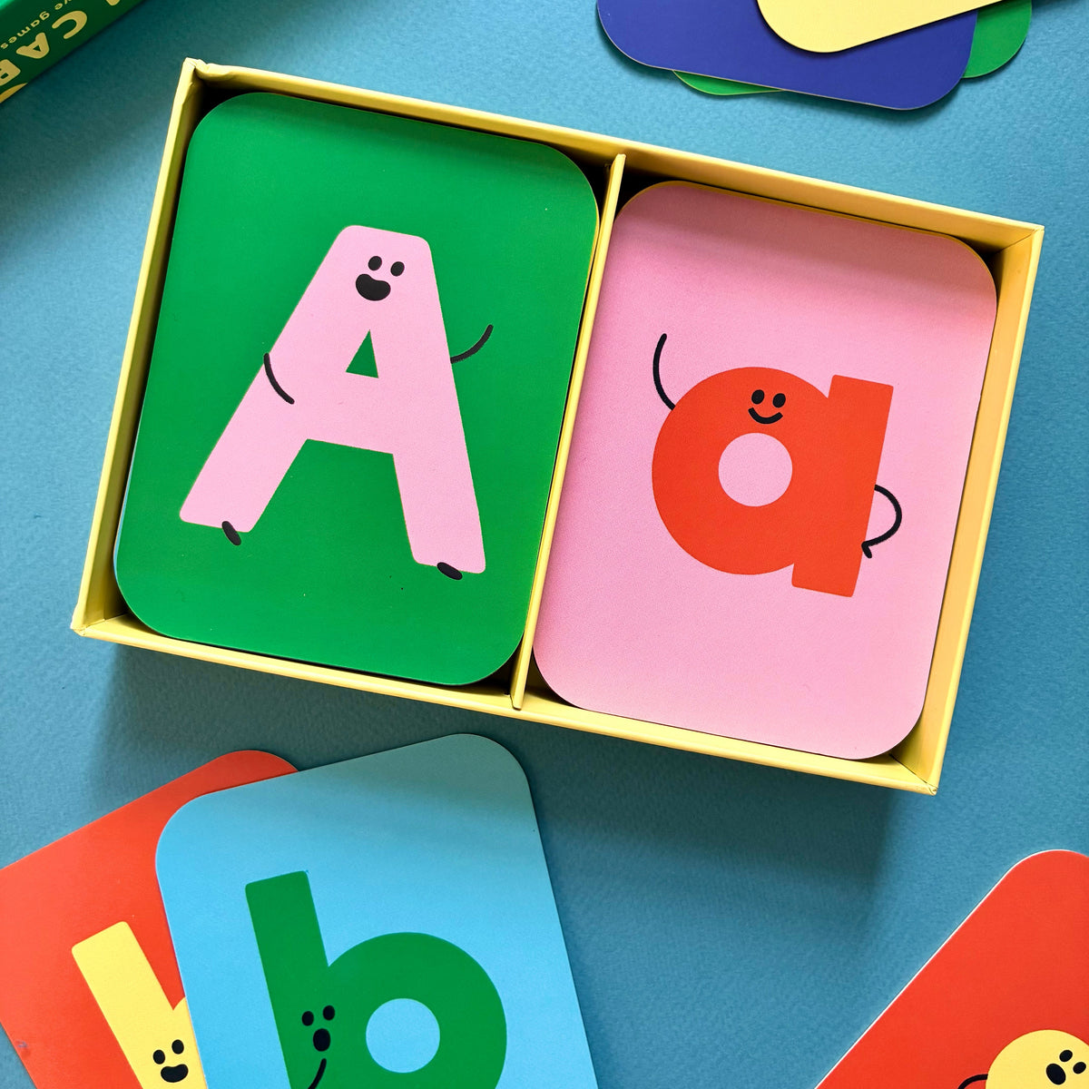 Close-up of uppercase A and lowercase a flashcards in an open yellow box, with more letter cards on a blue surface.